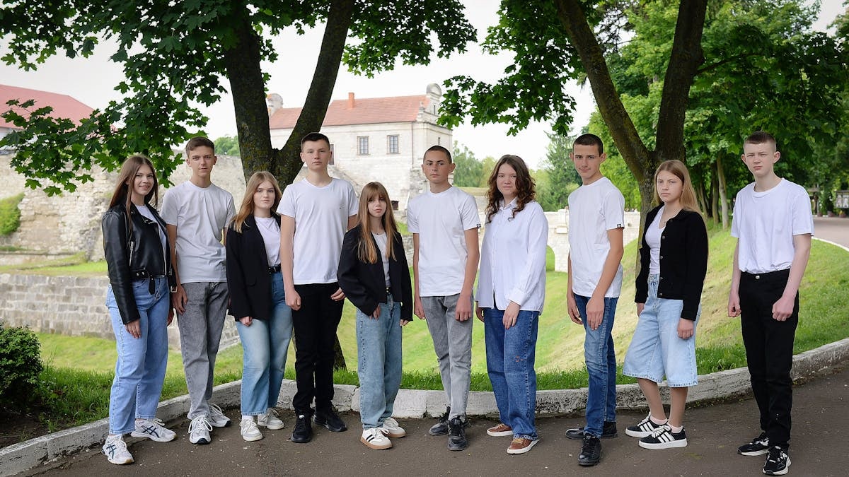 Group of teenagers in casual attire standing under trees in a park setting with historic building in background.
